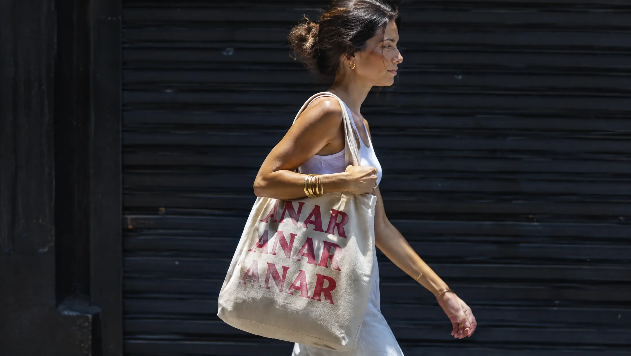 A woman walking past a dark shutter with an Anar canvas tote